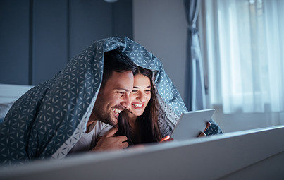 Couple dans une chambre allongé à plat ventre sur leur lit, la couette remontée sur leur tête, regardant un contenu sur leur tablette.